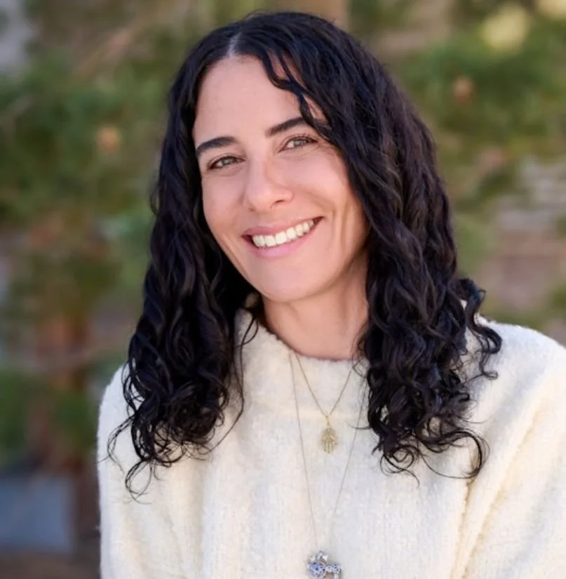 A woman with curly black hair smiling outdoors, wearing a white sweater and layered necklaces.