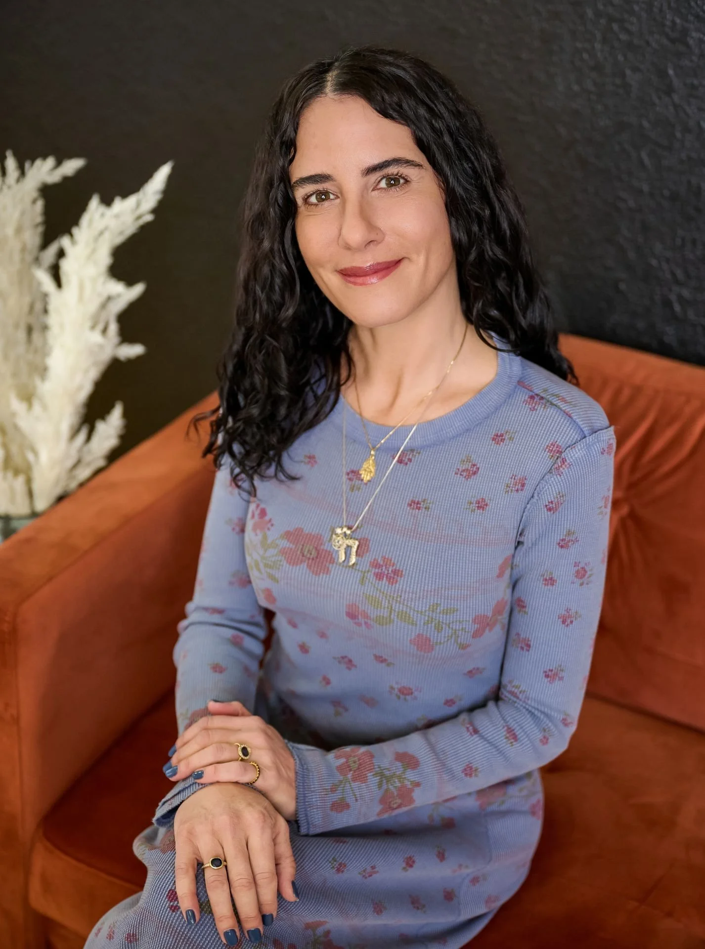A woman with dark, curly hair sitting on a brown sofa, wearing a blue floral dress, gold jewelry, and posing with her hands clasped.
