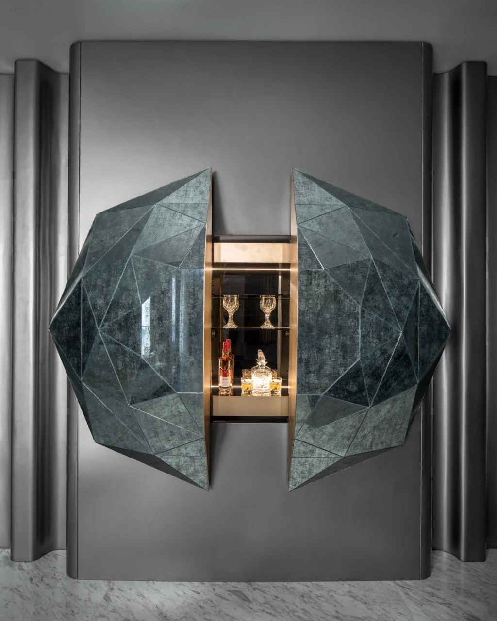 A modern decorative cabinet with a geometric, faceted glass front that opens to reveal a display of bottles and glasses inside.