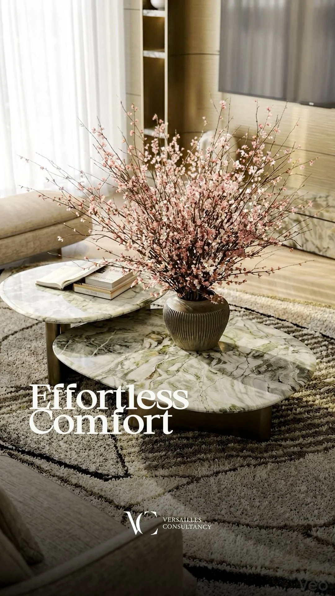 A living room with marble coffee tables, a beige sofa, a patterned area rug, a large pink floral arrangement in a gray vase, and a bookshelf in the background. Text reading "Effortless Comfort" and a logo for Versailles Consultancy are overlaid.