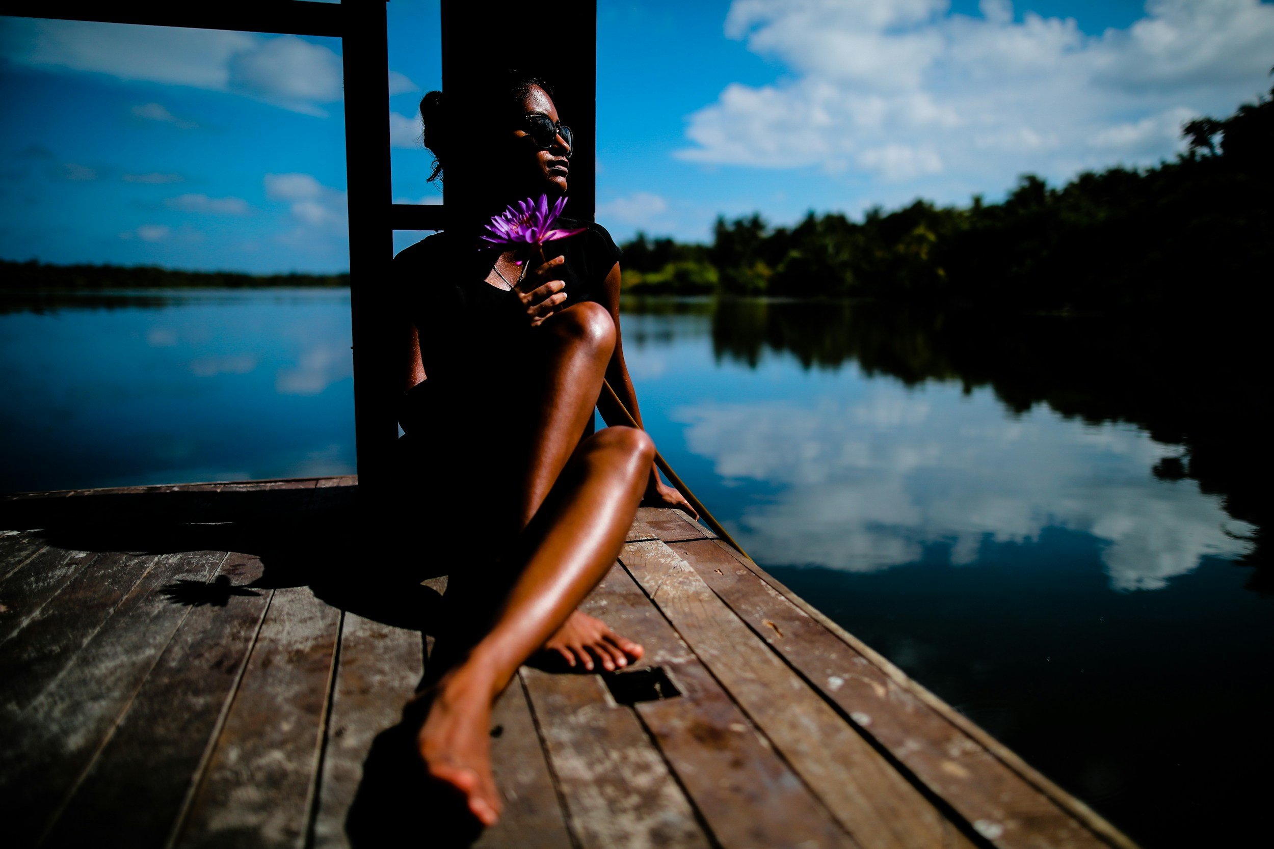 A woman sitting on a wooden dock by a lake, holding a purple water lily, wearing sunglasses, with trees and a cloudy sky reflected on the water.