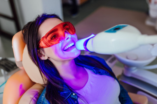 Woman undergoing teeth whitening treatment in a dental chair with UV light device, wearing protective red glasses and smiling.