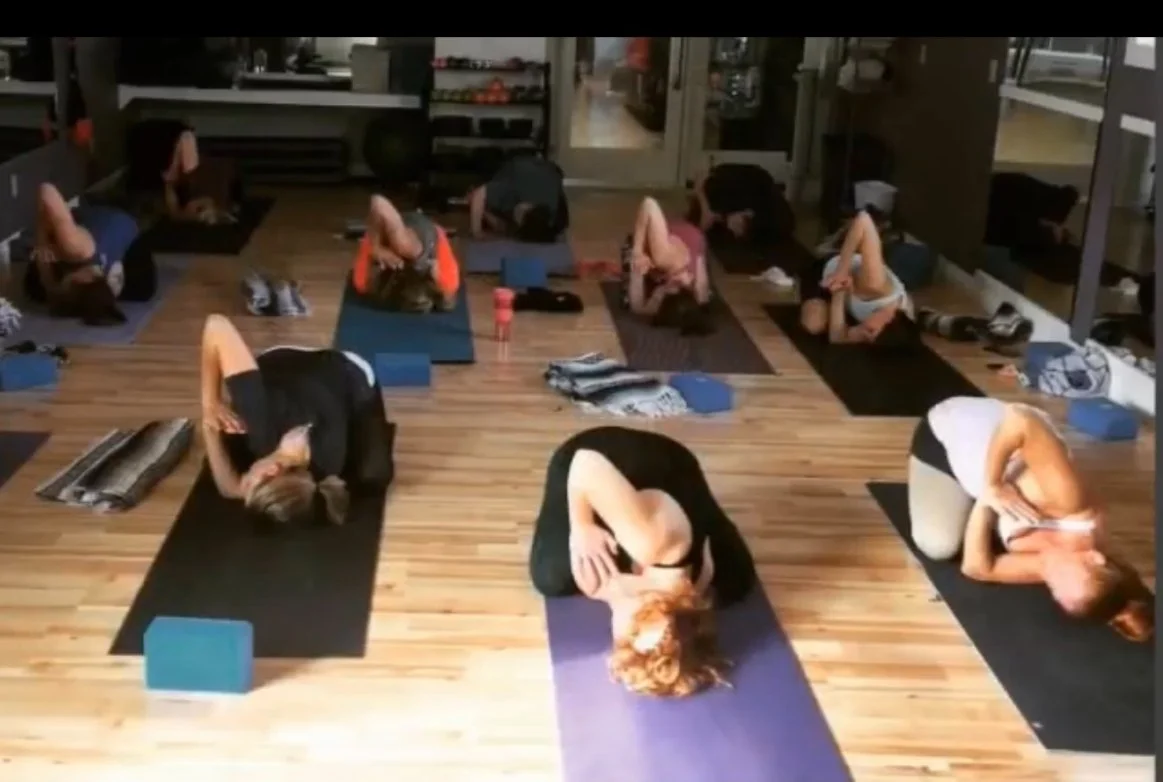 People participating in a yoga class in a studio with wooden floors, practicing seated forward bends.