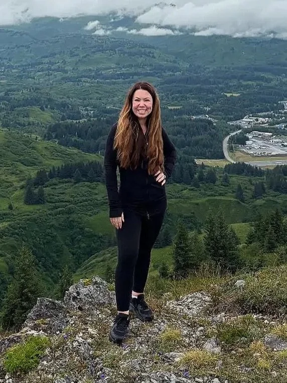 A woman in black athletic clothing standing on a rocky hillside with a green valley and mountains in the background.