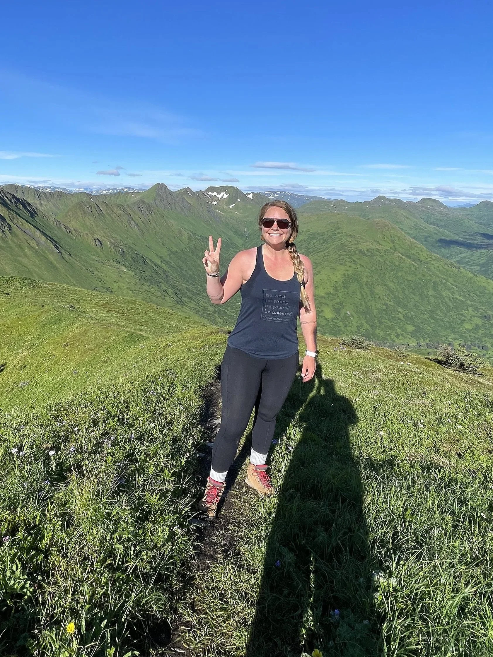 A woman smiling and making a peace sign with her right hand, standing on a grassy mountain trail with green mountains and a blue sky in the background.