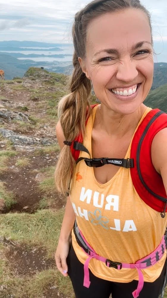 Woman with long braided hair smiling, wearing a yellow tank top with a red backpack and a pink waist strap, standing outdoors on a mountain trail with cloudy sky and hills in the background.
