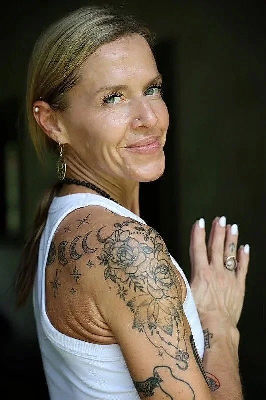 Close-up of a smiling woman with blonde hair, tattoos on her arm, wearing a white tank top, earrings, and a necklace, with her hand resting on her shoulder.