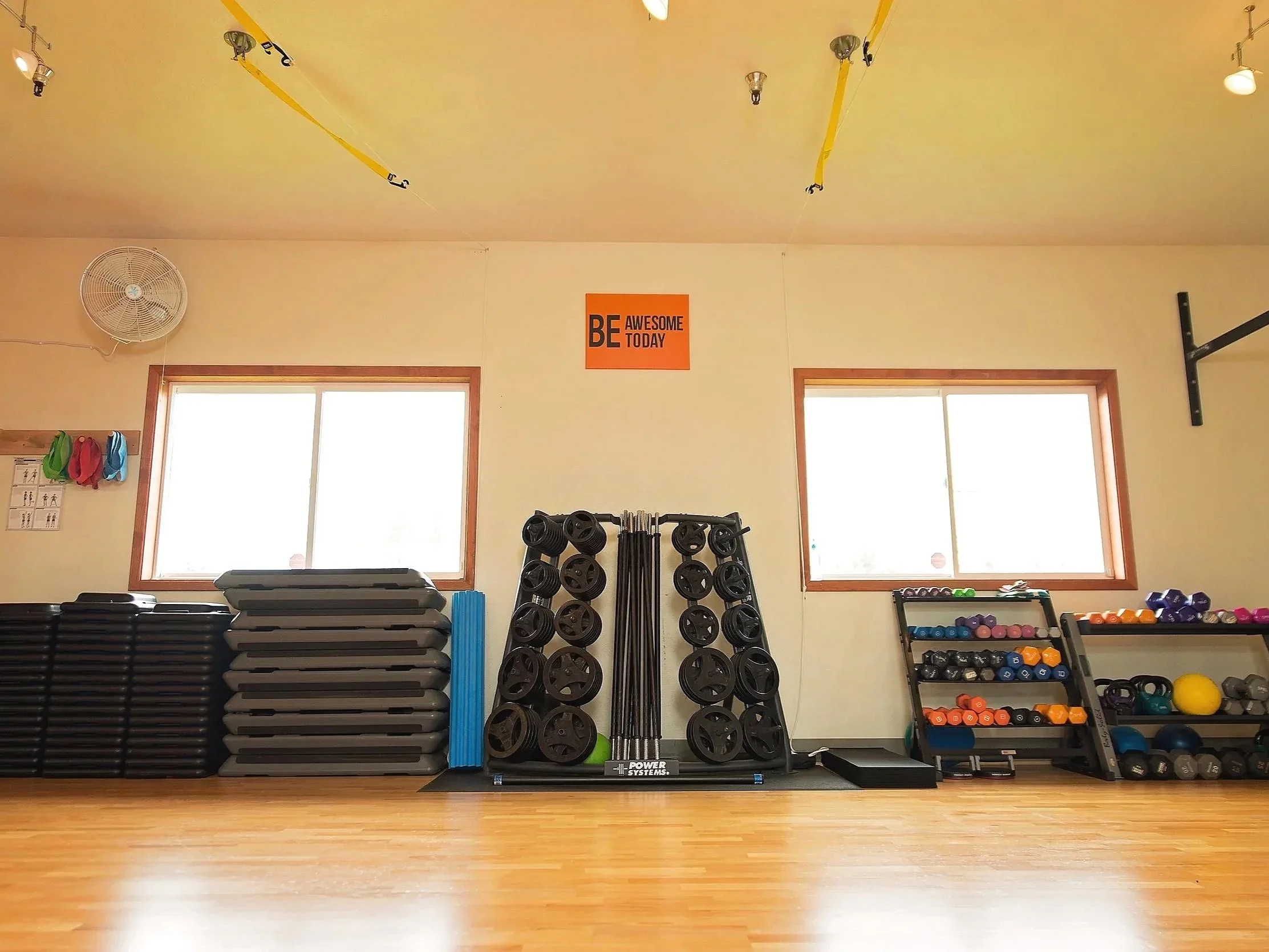 Empty gym room with fitness equipment and storage racks, two large windows, and a motivational sign on the wall that says "BE AWESOME TODAY."