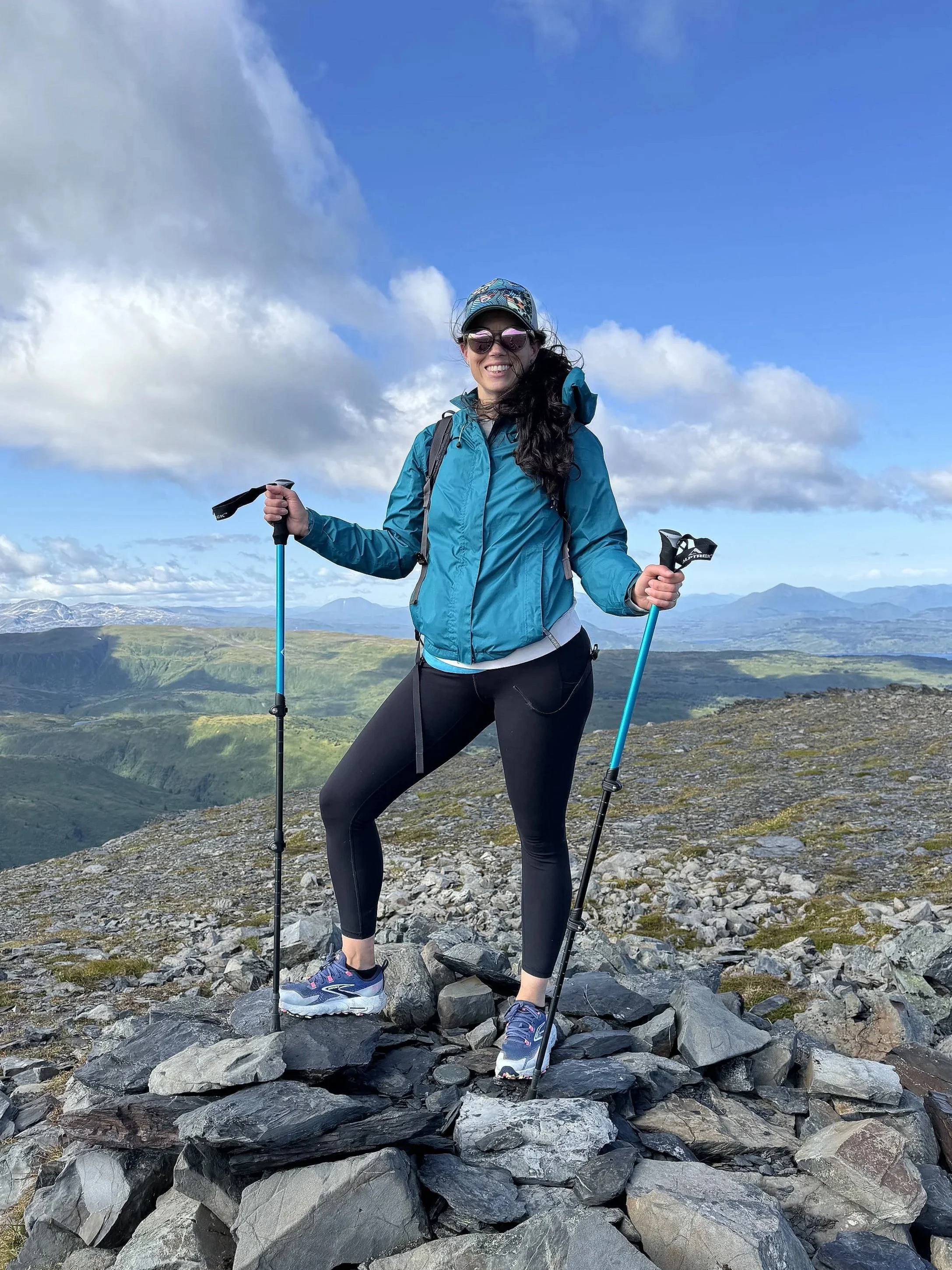 Woman standing on a rocky mountaintop with trekking poles, wearing outdoor gear, smiling, with green valleys and mountains in the background under a blue sky with clouds.