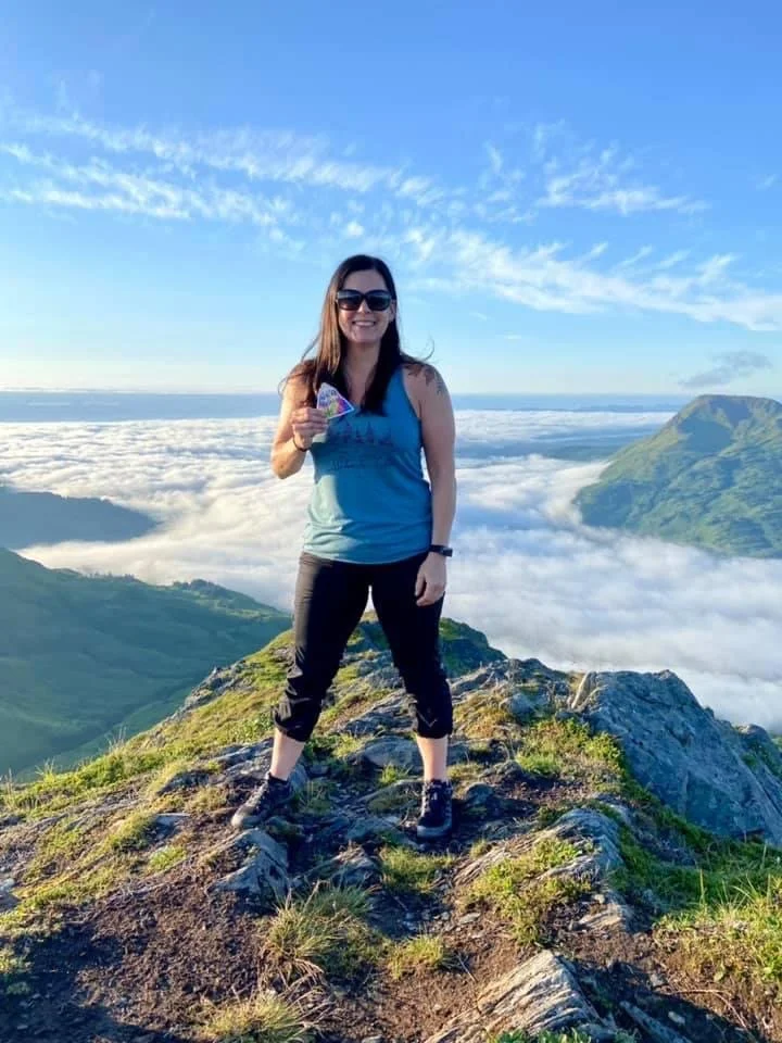 A woman smiling and holding a Change Your Altitude sticker stands on a mountain peak with a scenic backdrop of clouds, mountains, and blue sky in Kodiak, AK.