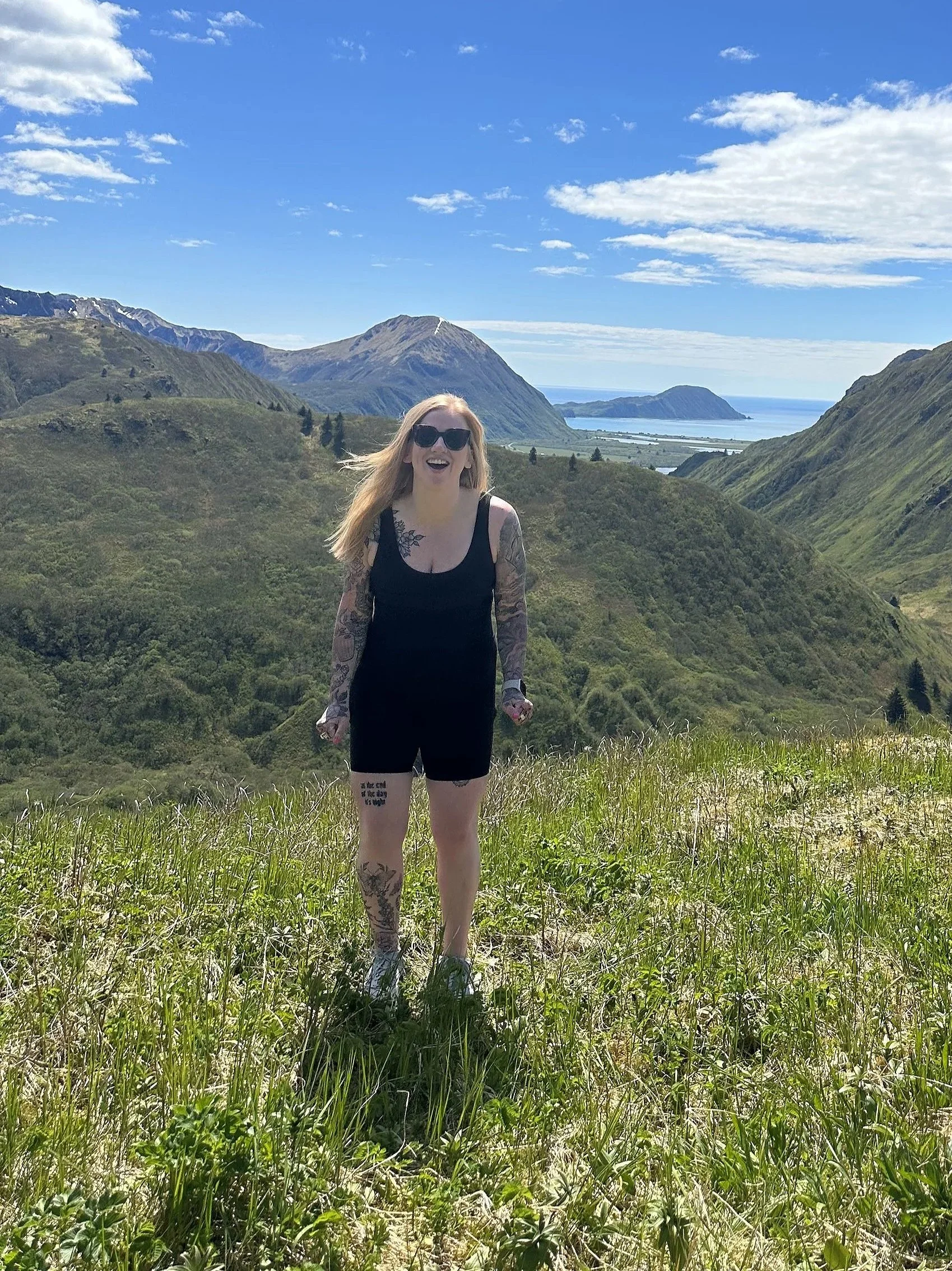 A woman with long blond hair, sunglasses, and tattoos on her arms and leg, smiling and standing on a grassy hill with a scenic backdrop of green mountains, a body of water, and a partly cloudy blue sky.