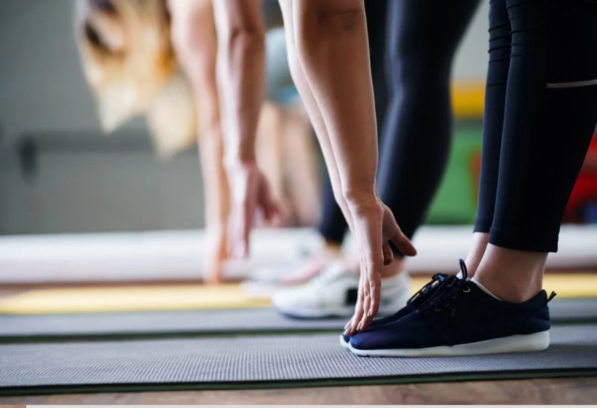 People doing yoga in a studio, standing on yoga mats, stretching downward with hands reaching toward the floor.