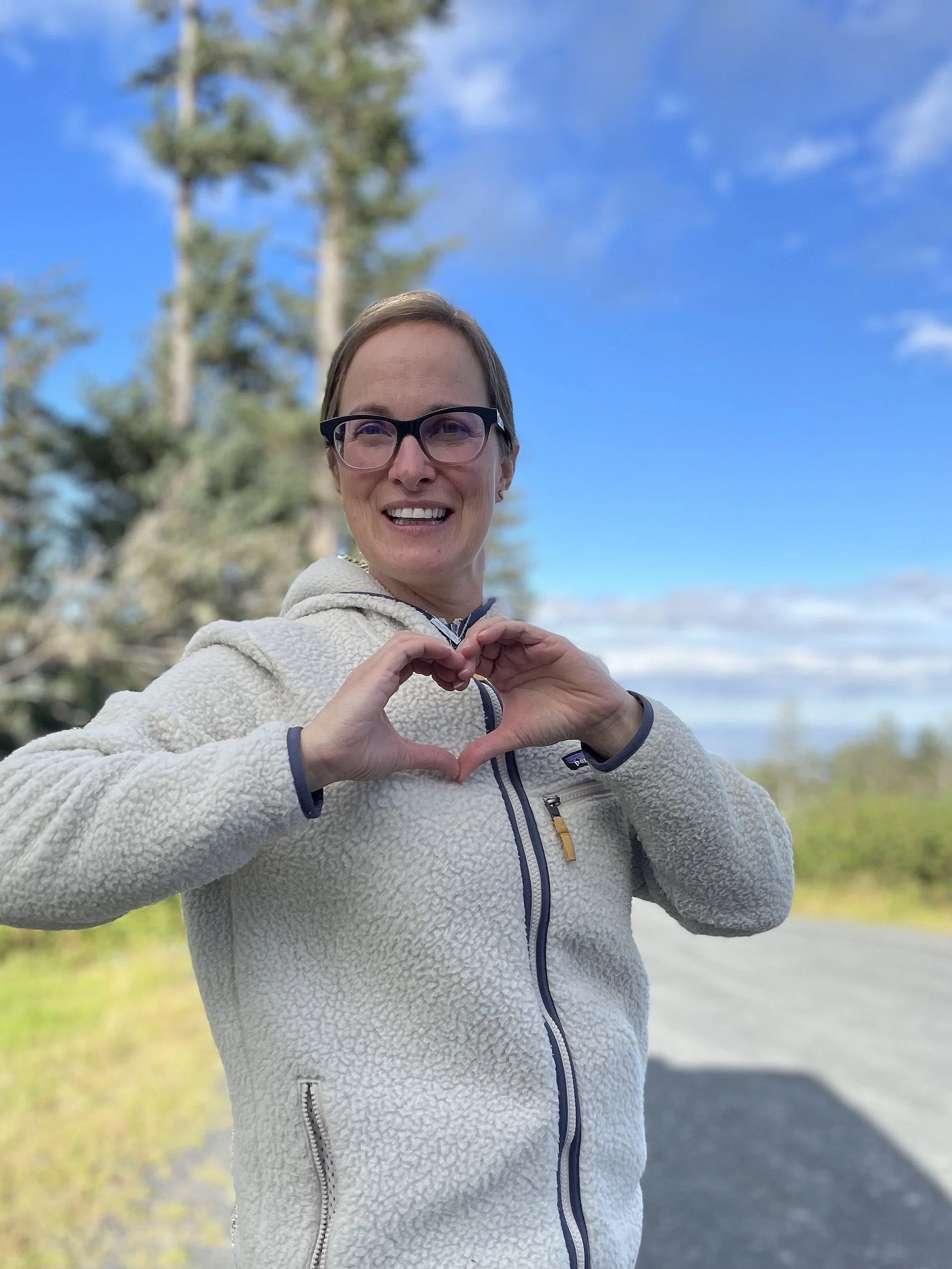A woman with glasses smiling and making a heart shape with her hands outdoors during the daytime, with a cloudy sky and trees in the background.