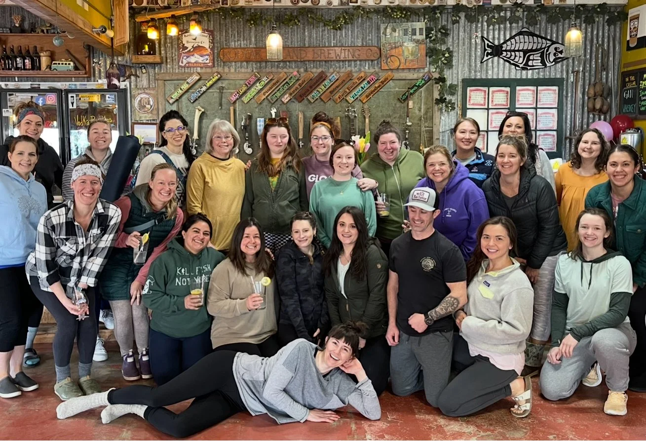 Group of smiling women and one man in casual attire posing inside a rustic bar or brewery, some holding drinks, with decorations on the wall including a fish sculpture and beer taps.