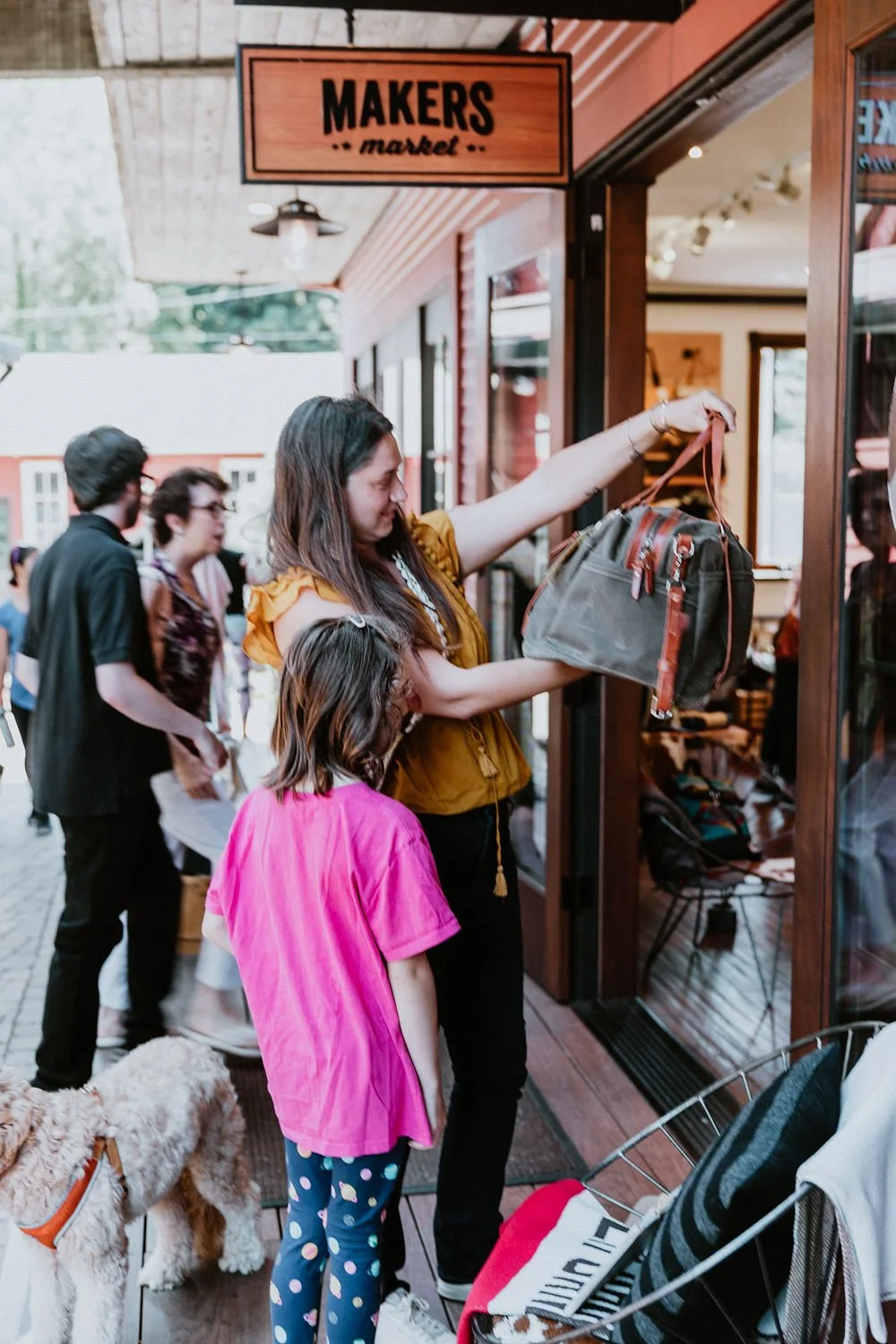 A woman and a young girl shopping outside a store called Makers Market, with a group of people and a dog nearby.