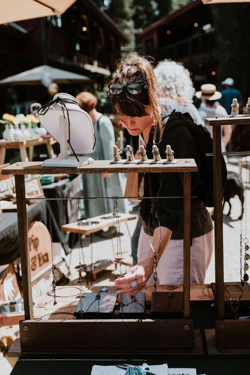 A woman with brown hair, glasses on her head, and wearing a black hoodie is browsing jewelry at an outdoor market stall. The stall displays necklaces, bracelets, and other jewelry on wooden racks and tables. Other shoppers are visible in the backgrou