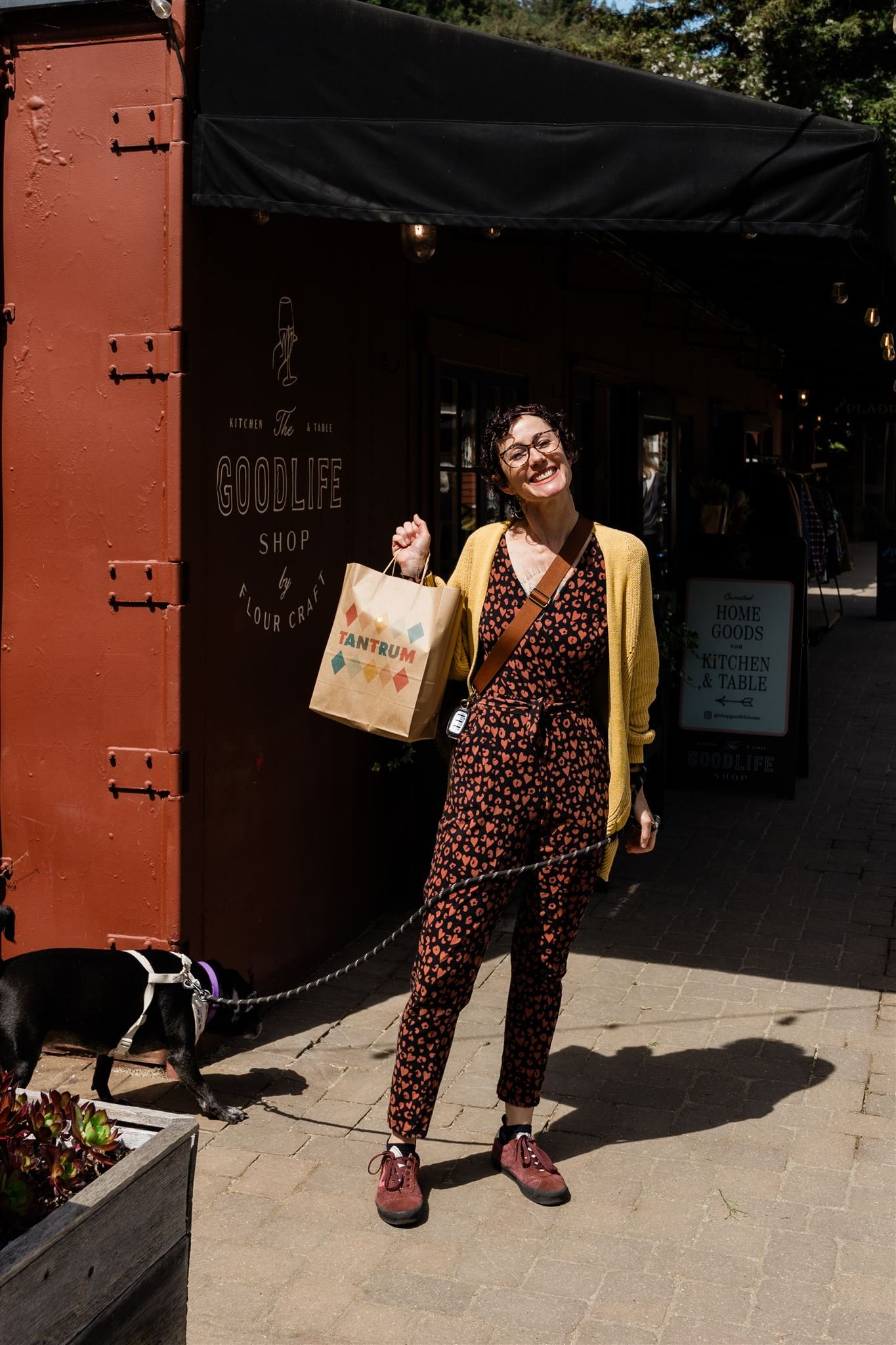 A woman with curly hair and glasses smiling and holding a shopping bag standing outside a store named 'Goodlife'. She is wearing a yellow cardigan over a floral jumpsuit and hiking boots. A black and white dog is on a leash near her, with its head do