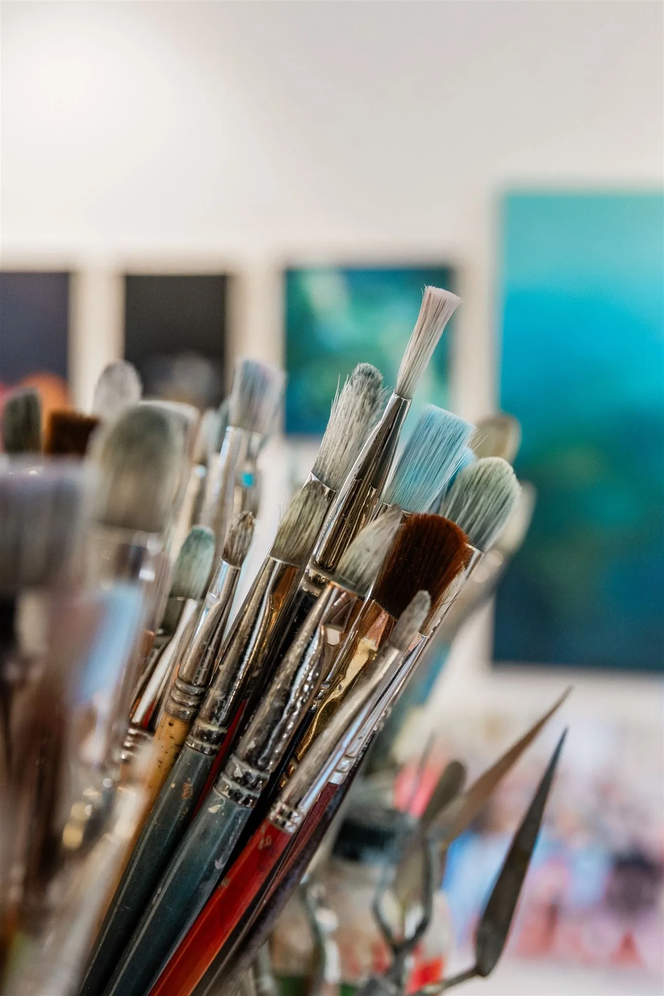 A collection of paintbrushes with various bristle shapes and sizes, standing upright in a container, with paintings hanging on the wall in the background.