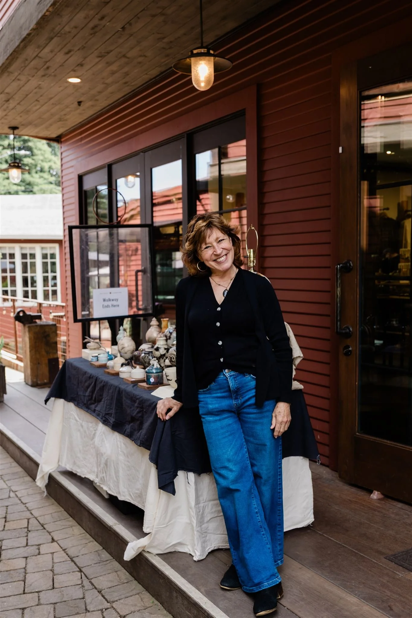 A smiling woman with short, wavy brown hair, wearing a black jacket and blue jeans, standing outside a building with red wooden siding and large windows. A table with various ceramics and decorative items is visible behind her, and there are hanging 