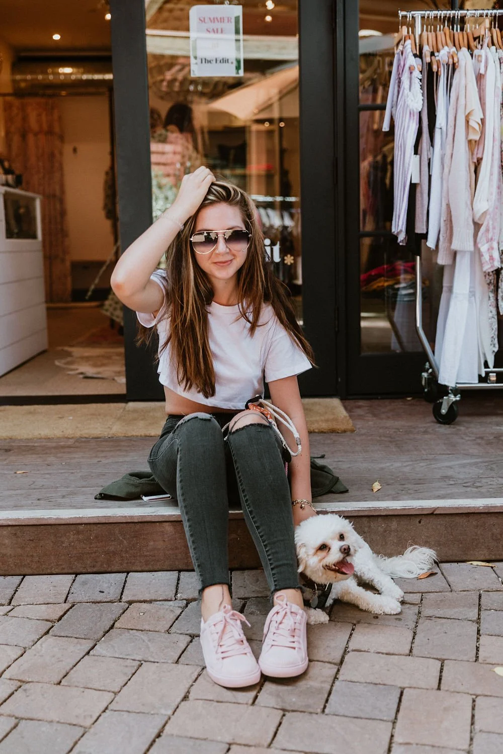 A young woman with long brown hair, wearing sunglasses, a white t-shirt, ripped black jeans, and pink sneakers, sitting on the curb outside a clothing boutique. She is smiling and touching her hair, with a small white fluffy dog laying beside her, to