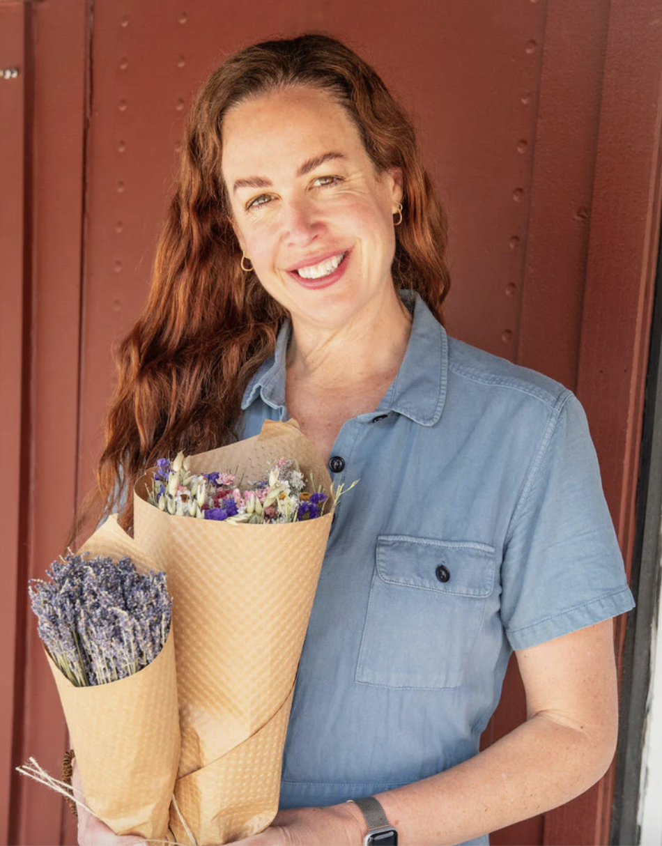 A woman with long red hair smiling and holding two bouquets of lavender and other wildflowers wrapped in brown paper, standing in front of a reddish-brown wall.