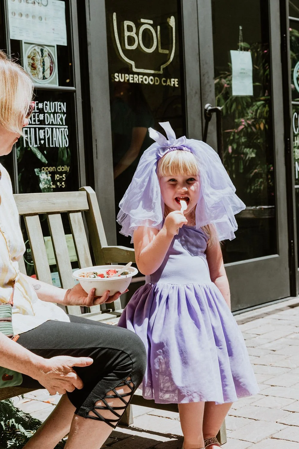 A young girl in a lavender dress and veil is eating ice cream cone outside a cafe, sitting on a bench next to a woman holding a bowl of food.