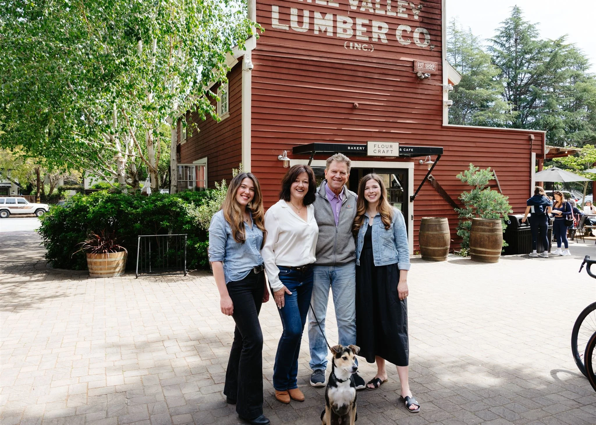 Group of four people smiling and standing outdoors in front of a red wooden building with a sign reading Bakery, Flour, Craft, Café. Two women and one man, with one woman holding a dog and a dog on a leash. Trees and parked cars in the background.