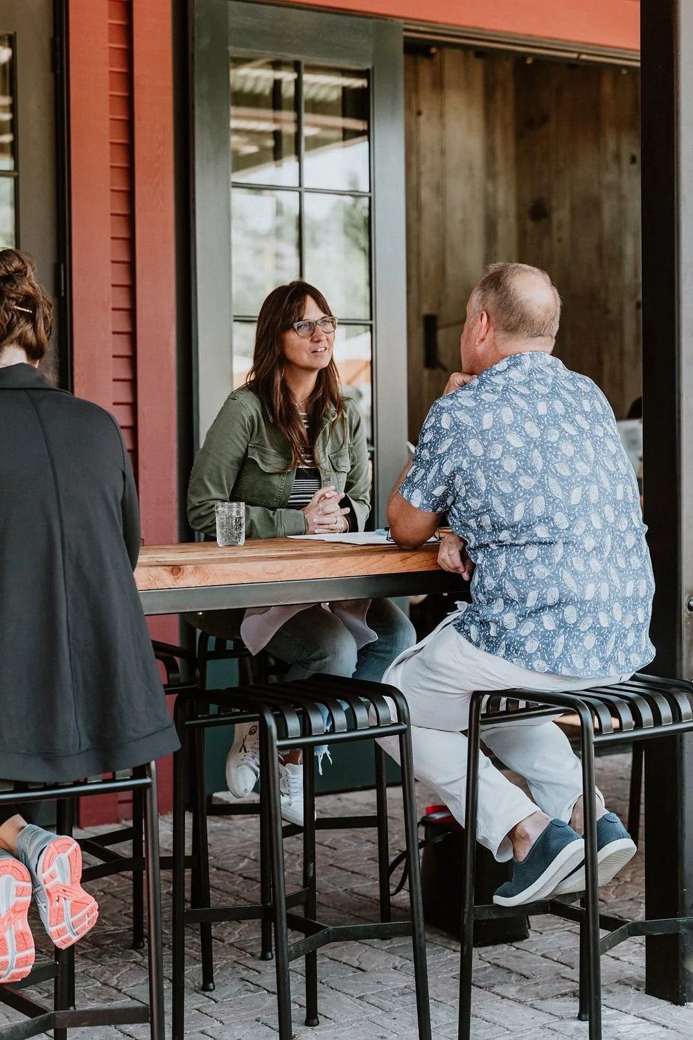 A woman with glasses and a green jacket sitting at an outdoor seating area, talking to a man in a patterned blue shirt, while another woman with a black jacket is partially visible.
