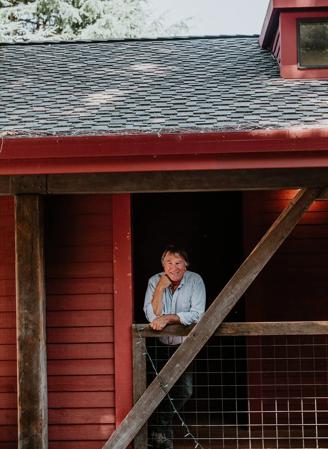 Smiling man leaning on a wooden railing in a rustic red house with a shingled roof and a small window.