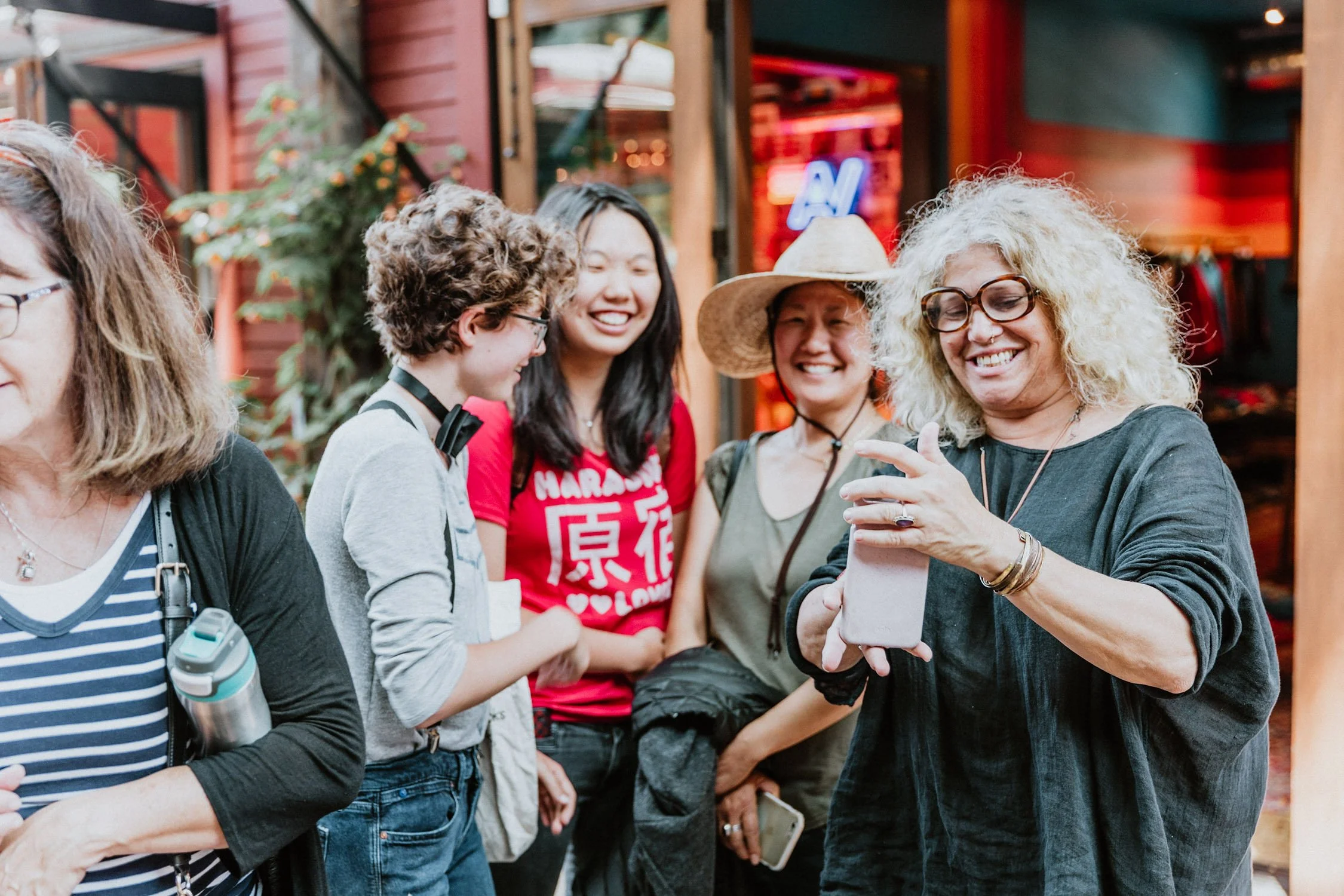 A group of women standing outdoors at a social event, looking at a smartphone together and smiling.