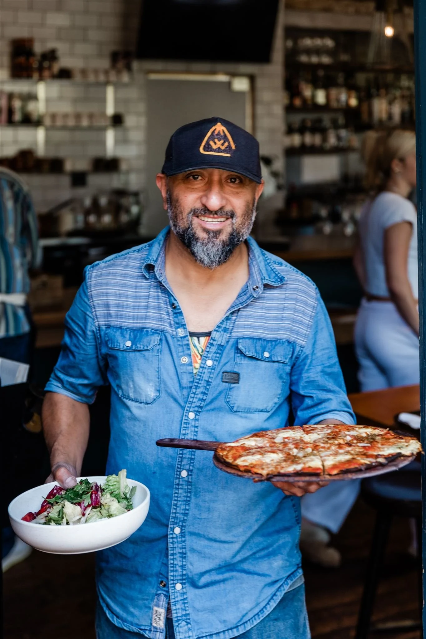 A smiling man with a beard wearing a blue shirt and a black cap, holding a bowl of salad in one hand and a pizza in the other inside a restaurant. There is a woman blurred in the background.
