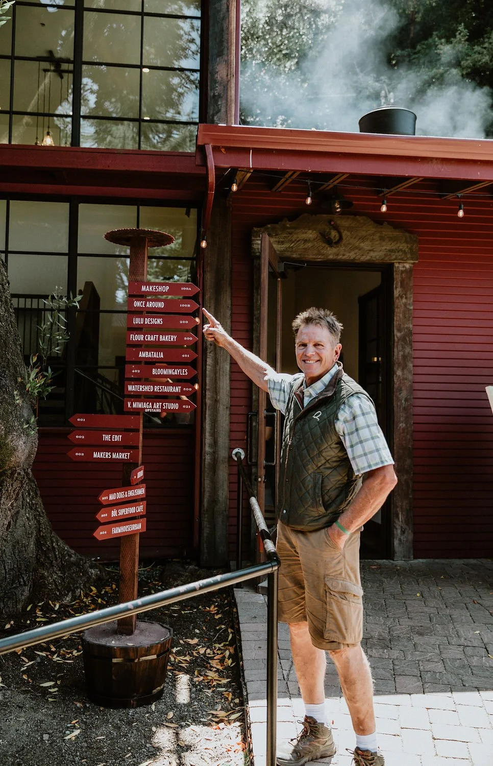 A man smiling and pointing at a red directional sign outside a building with large windows, surrounded by trees.