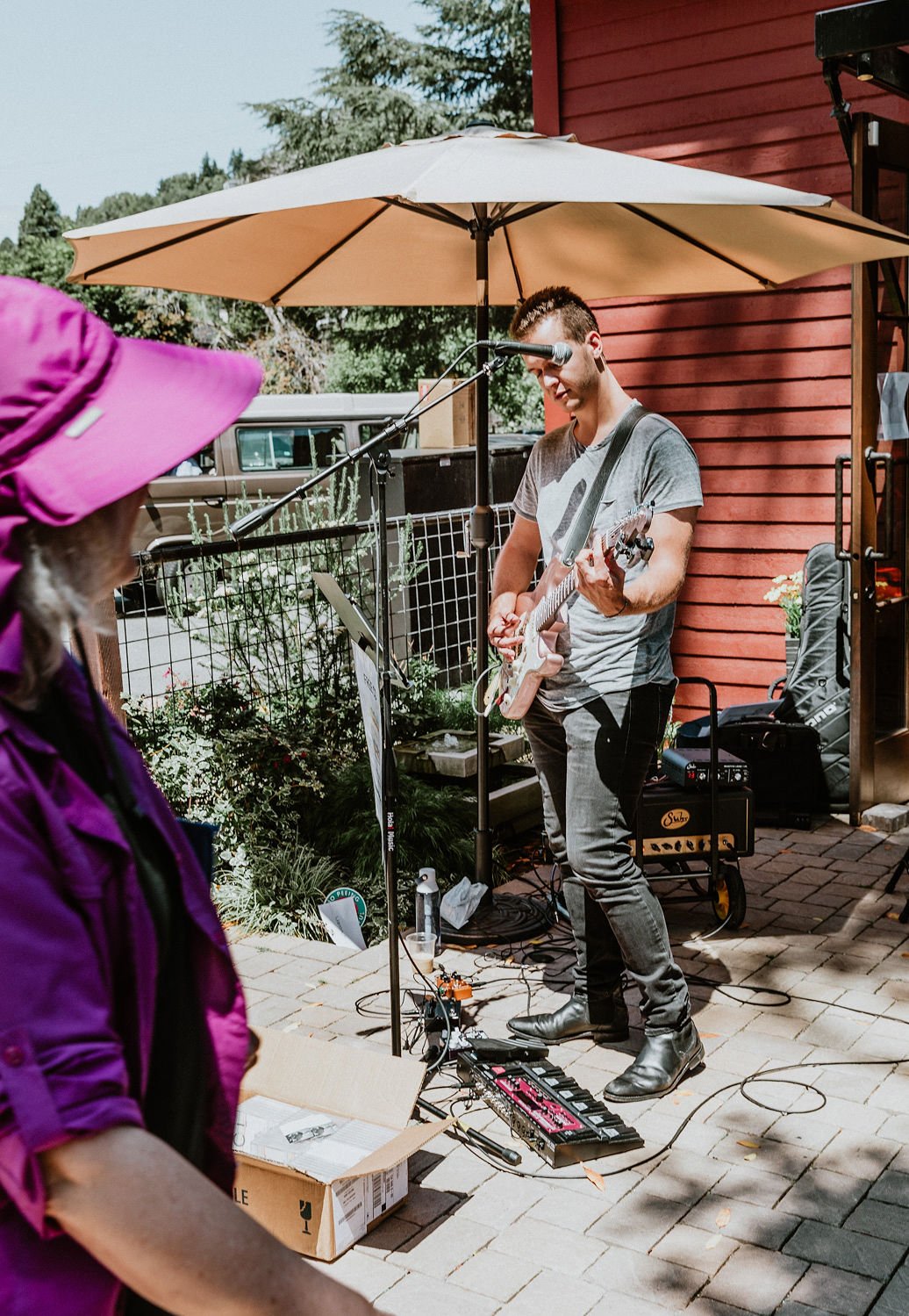 A man playing an electric guitar and singing into a microphone outdoors under a large umbrella, with an audience member in a purple hat watching.