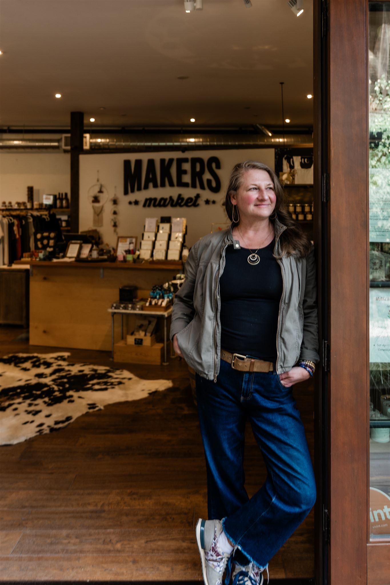 A woman stands in the doorway of Makers Market, smiling and looking to her right, inside a retail store with various products on display and a cowhide rug on the wooden floor.