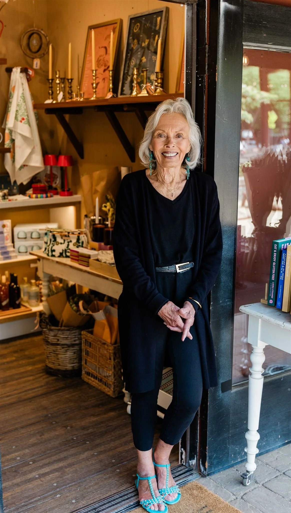 An elderly woman with gray hair smiling, standing by a storefront entrance, wearing a black outfit and turquoise high-heeled sandals, inside the shop with shelves of decorative items and books.