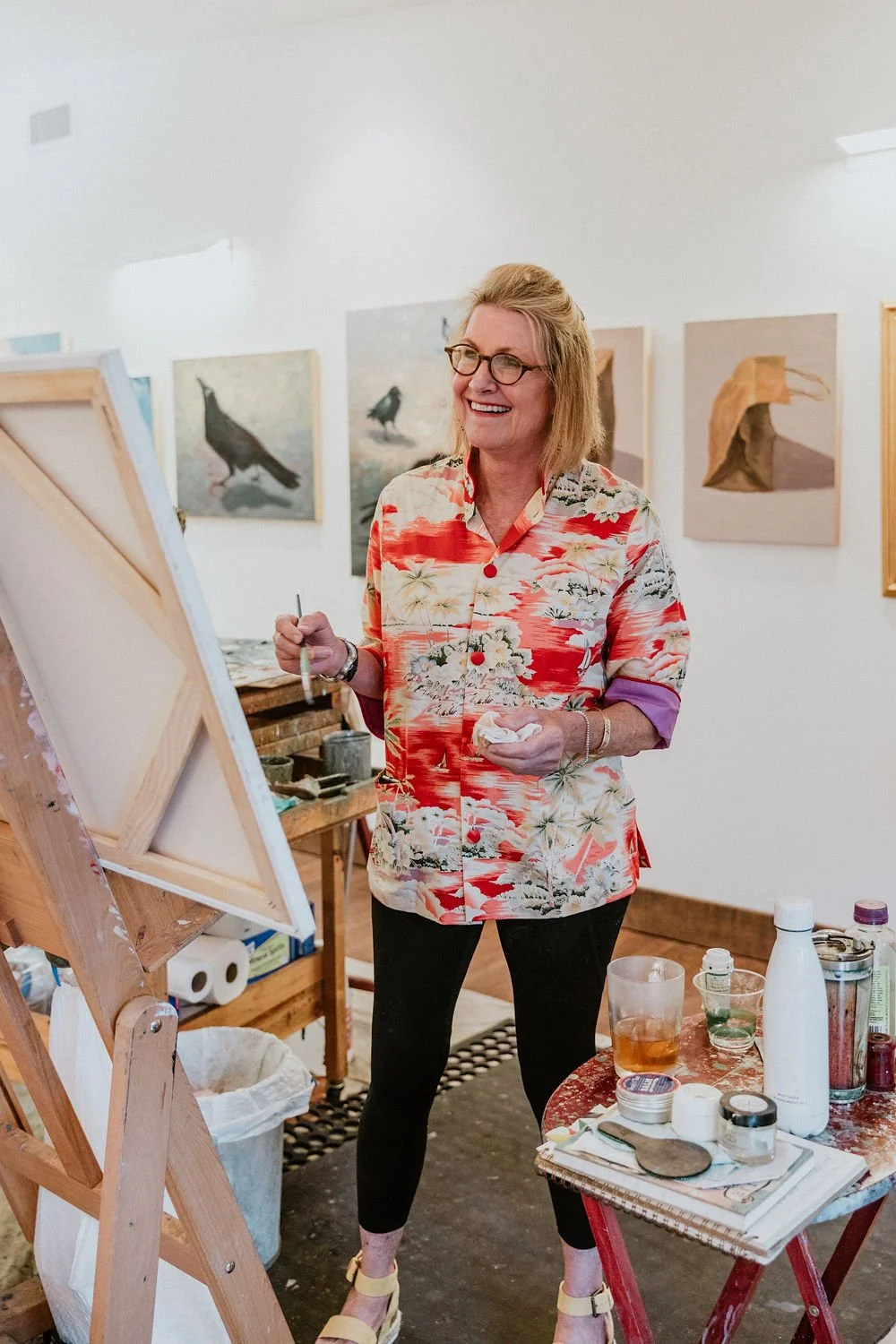 A woman with blonde hair and glasses painting on an easel in an art gallery, wearing a colorful floral shirt, with paintings of birds hanging on the white wall behind her, and art supplies on a table nearby.