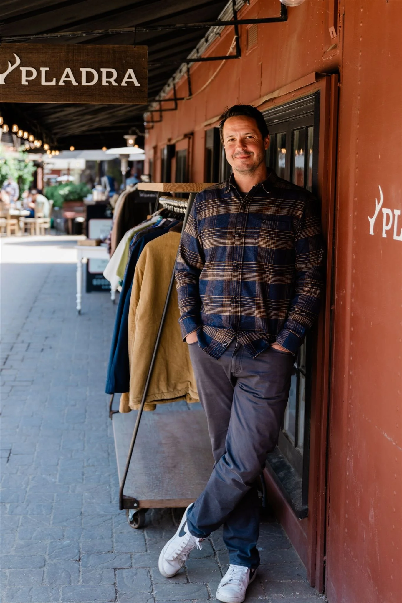 A man with dark hair and facial hair leaning against a red wall, smiling. He is wearing a dark, plaid shirt and gray pants, with white sneakers. Behind him is a clothing rack with jackets, and a street scene with outdoor seating and people, under str