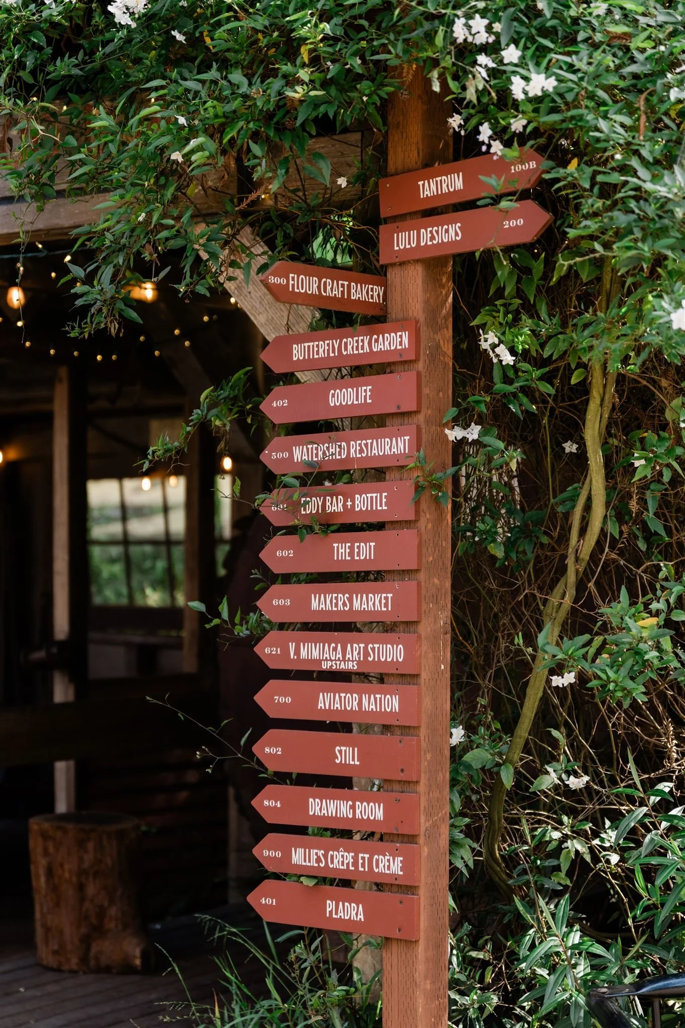 Wooden signpost with multiple directional signs surrounded by green leafy plants and white flowers. The signs indicate directions and distances to various locations such as Tantrum, Lulu Designs, Flour Craft Bakery, Butterfly Creek Garden, Goodlife, Watershed Restaurant, Eddy Bar + Bottle, The Edit, Makers Market, V. Mimiaga Art Studio, Aviator Nation, Still, DSW, Drawing Room, Millie’s Crêpe et Crème, and Plada.