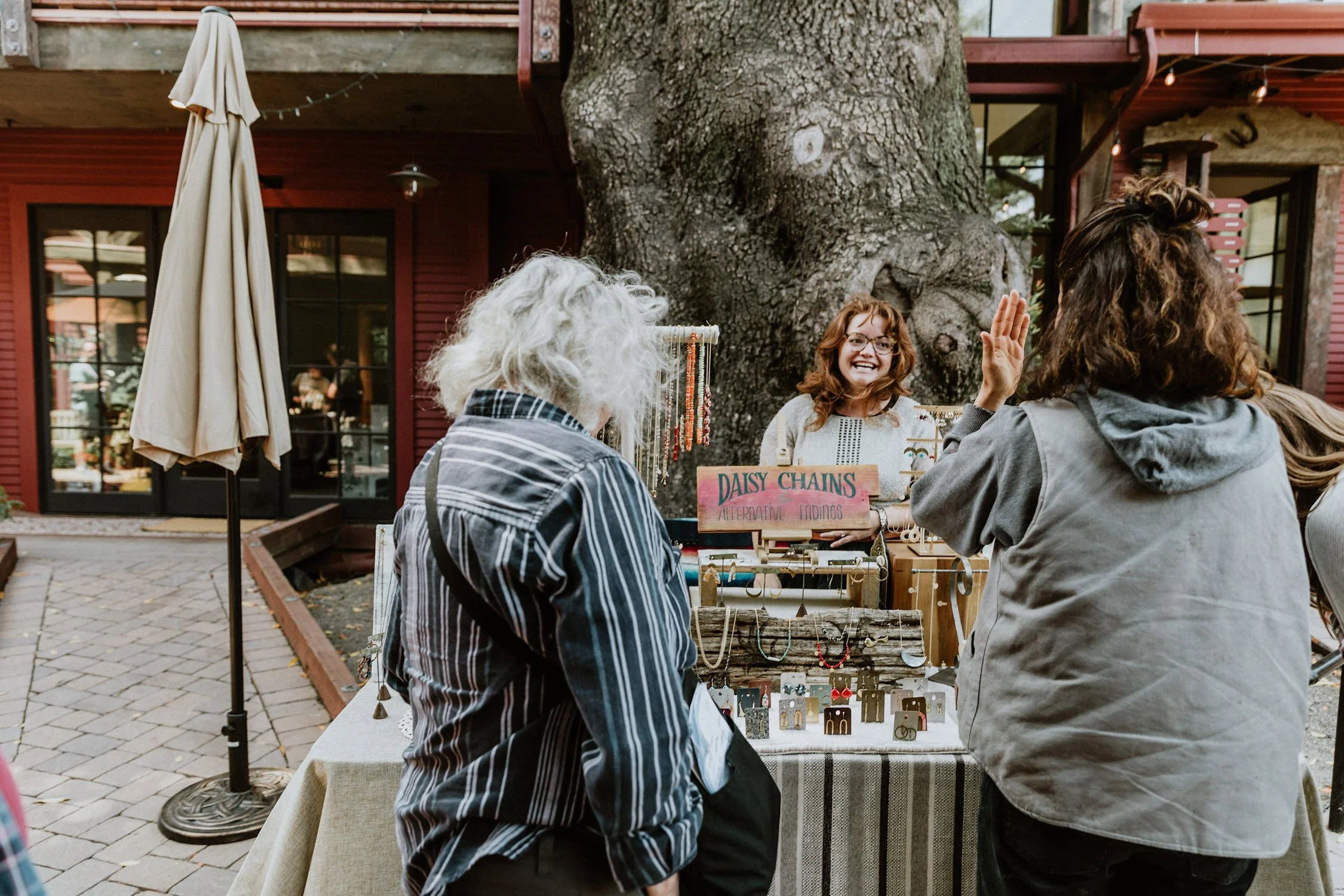 Two women and a young girl browsing jewelry at an outdoor market stall under a large tree, with a red building in the background.