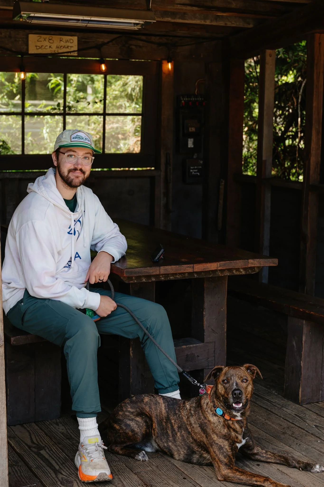 A man with glasses and a beard, wearing a cap, hoodie, and green pants, sitting at a wooden table with a grey retractable leash, and a brindle-colored dog lying on the wooden porch next to him. The porch is rustic with a large window and some visible