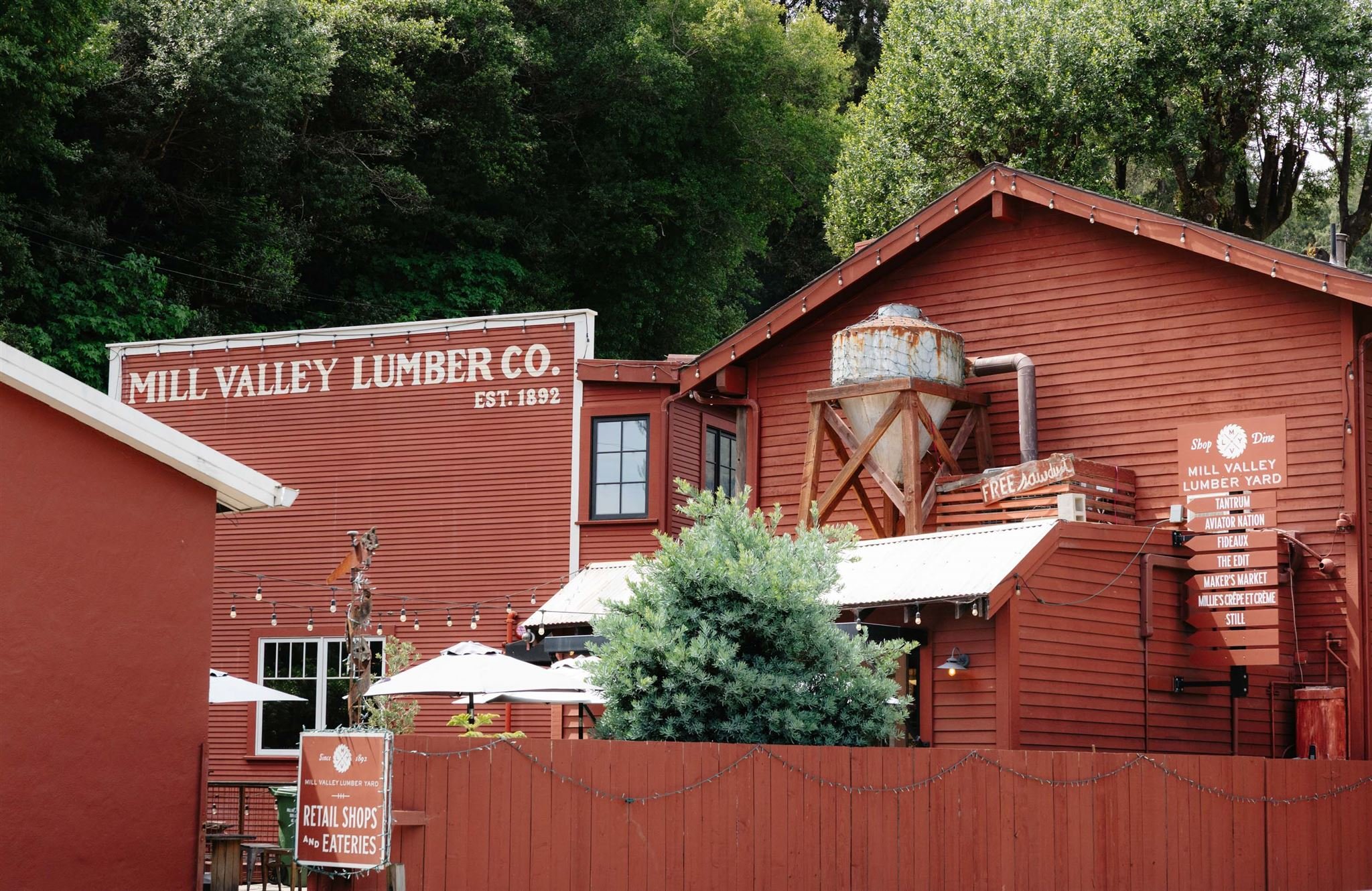 Red wooden building with signs indicating 'Mill Valley Lumber Co.', established in 1892, and additional signage pointing to retail shops and eateries. Surrounding trees and outdoor seating area with umbrellas are also visible.