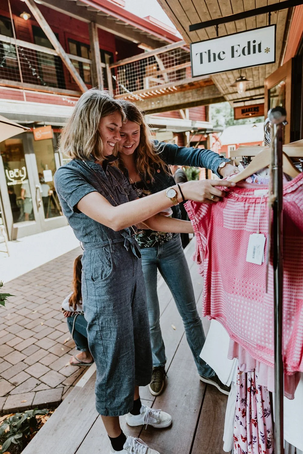 Two women shopping for clothes at an outdoor store, looking at a pink dress on a hanger and smiling.