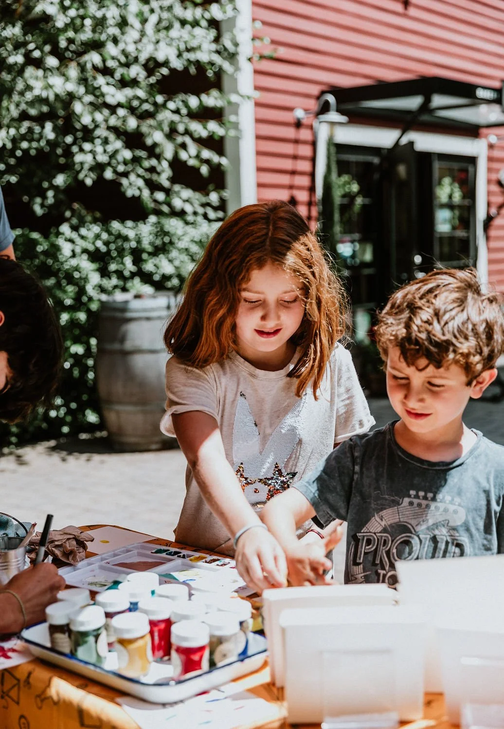 Children painting and drawing at an outdoor table with bottles of paint and art supplies, with a backyard and red building in the background.