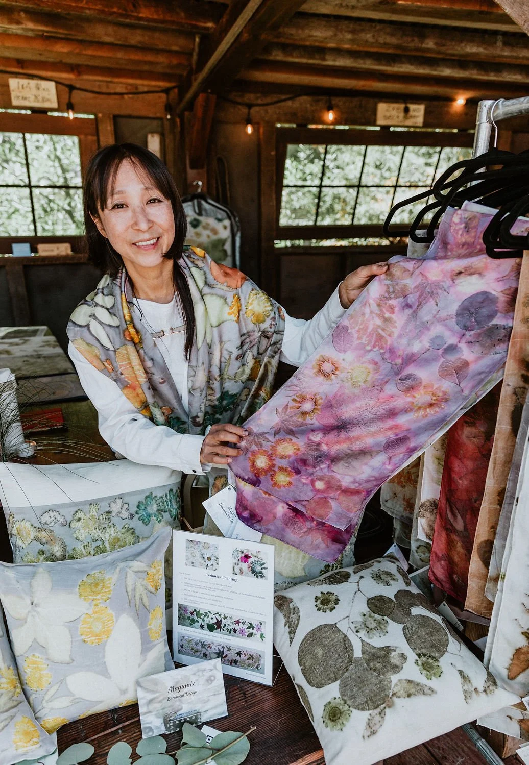 A woman standing inside a rustic, wooden room, holding a pink and purple floral fabric sample. She is smiling and dressed in a colorful scarf and white shirt, surrounded by botanical-themed pillows and fabric swatches on display.
