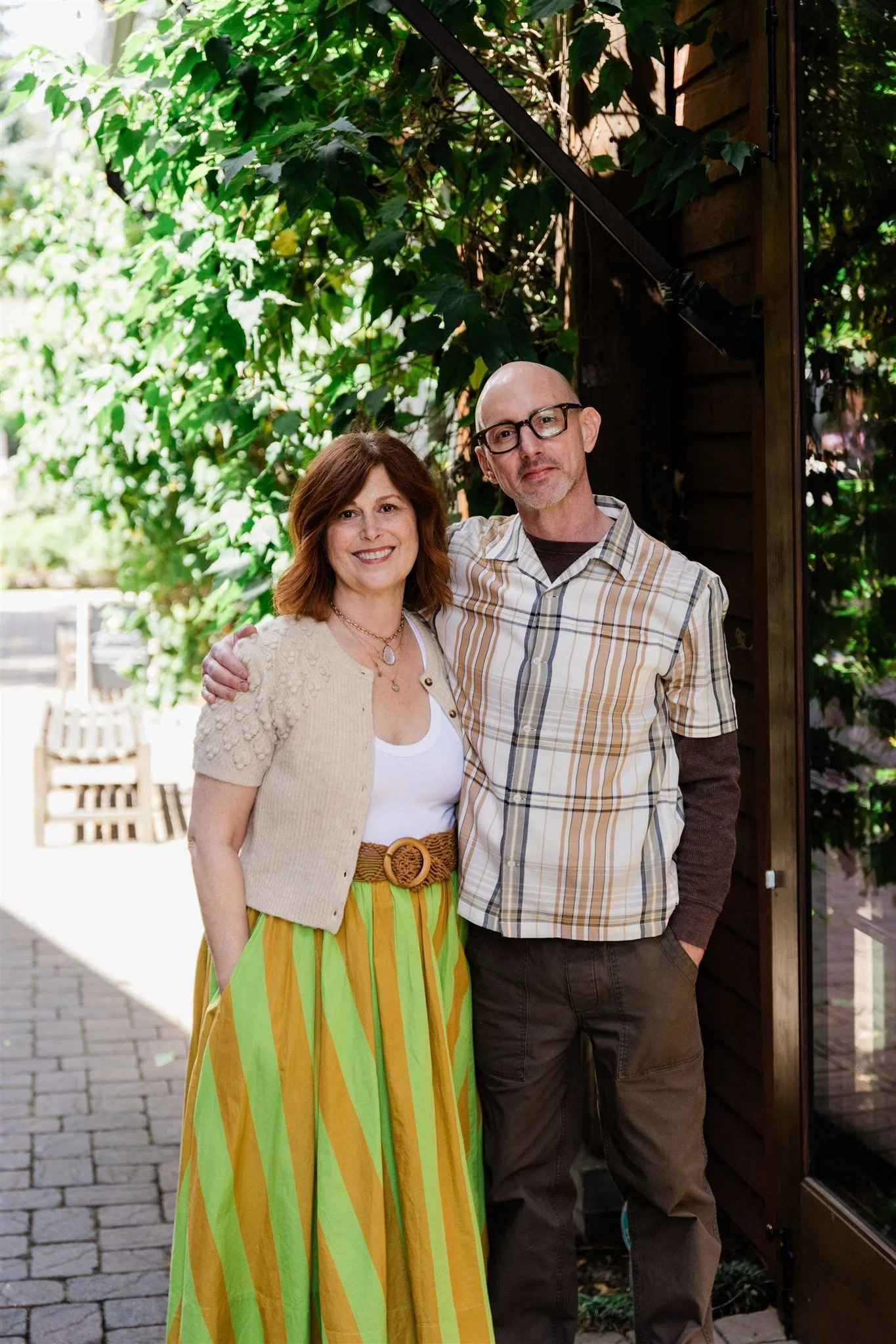 A man and woman standing close together outside, smiling, with foliage and a wooden wall behind them.