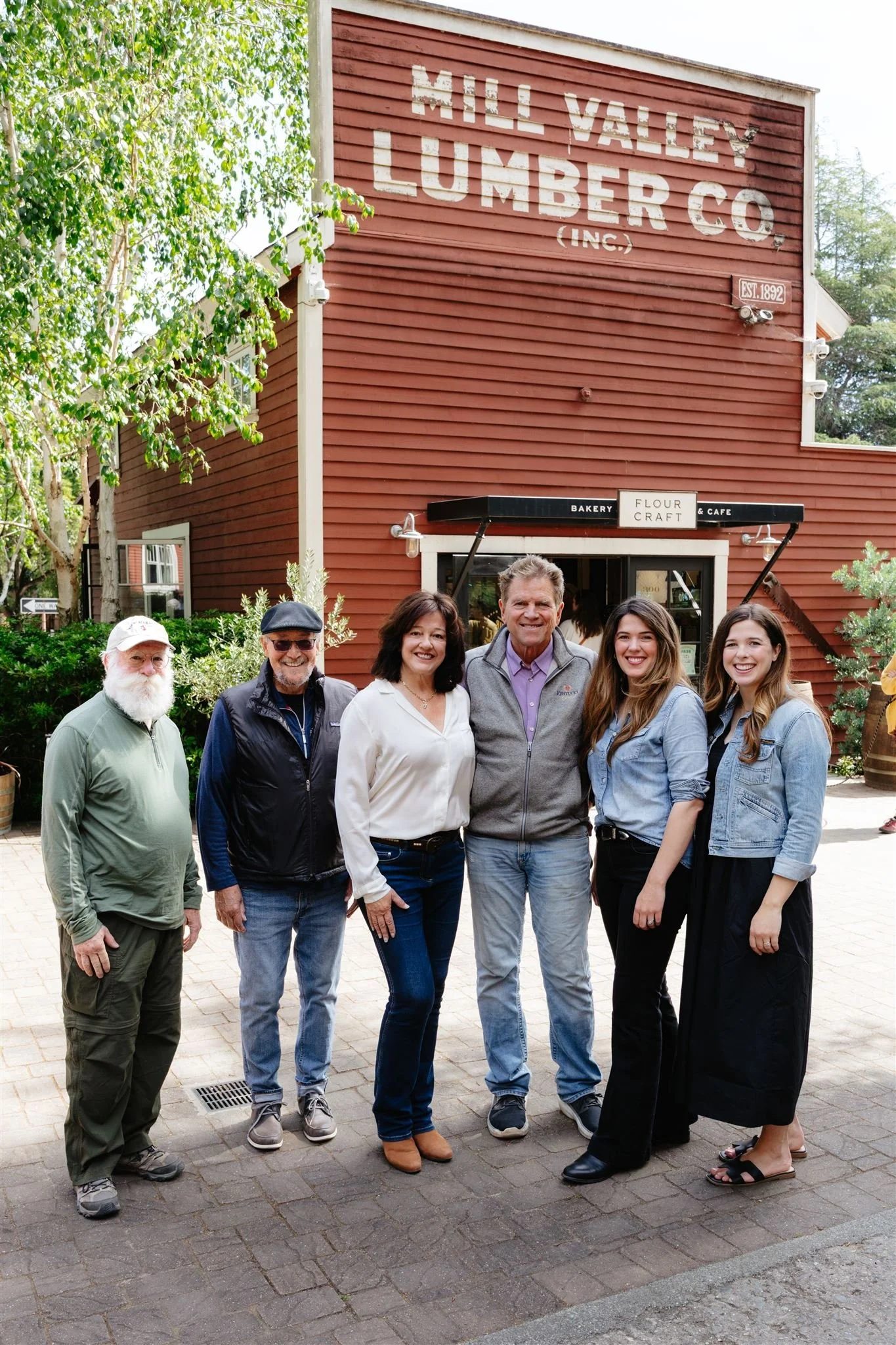 Group of six people standing outside in front of a red building with a sign that reads 'Mill Valley Lumber Co' and a bakery sign that says 'Flour, Craft & Cafe.' The group is smiling and dressed casually.