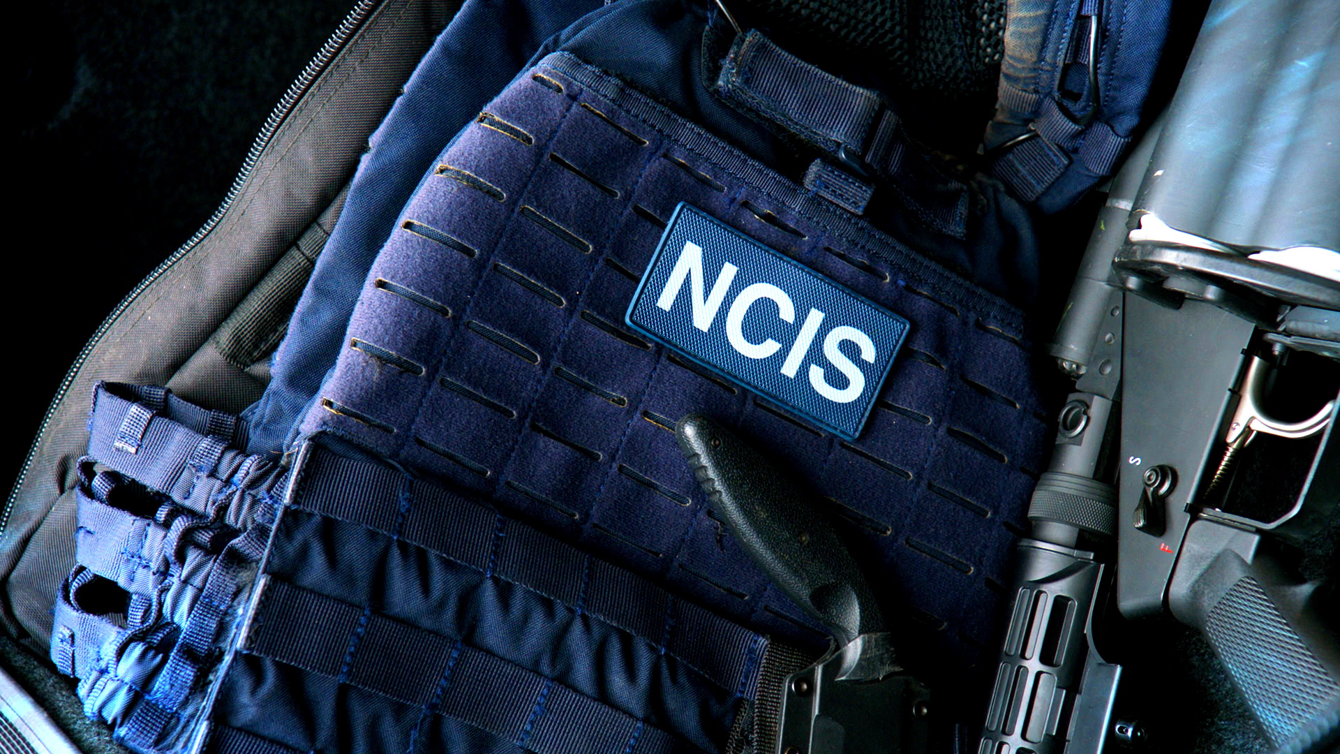 Close-up of a dark blue tactical backpack with an NCIS patch, a black rubber handle, and a firearm next to it.