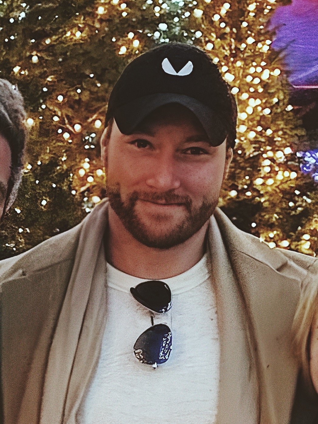 A man with a beard wearing a black cap and sunglasses hanging on his white shirt, standing in front of a tree with golden lights.