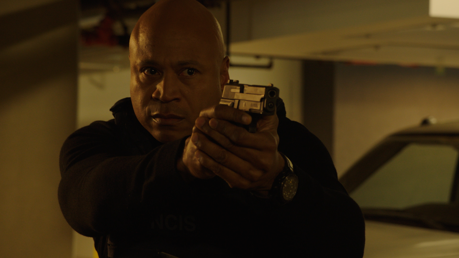 A man with a bald head and dark skin aiming a gun, wearing a black jacket and a watch, in an indoor parking garage.