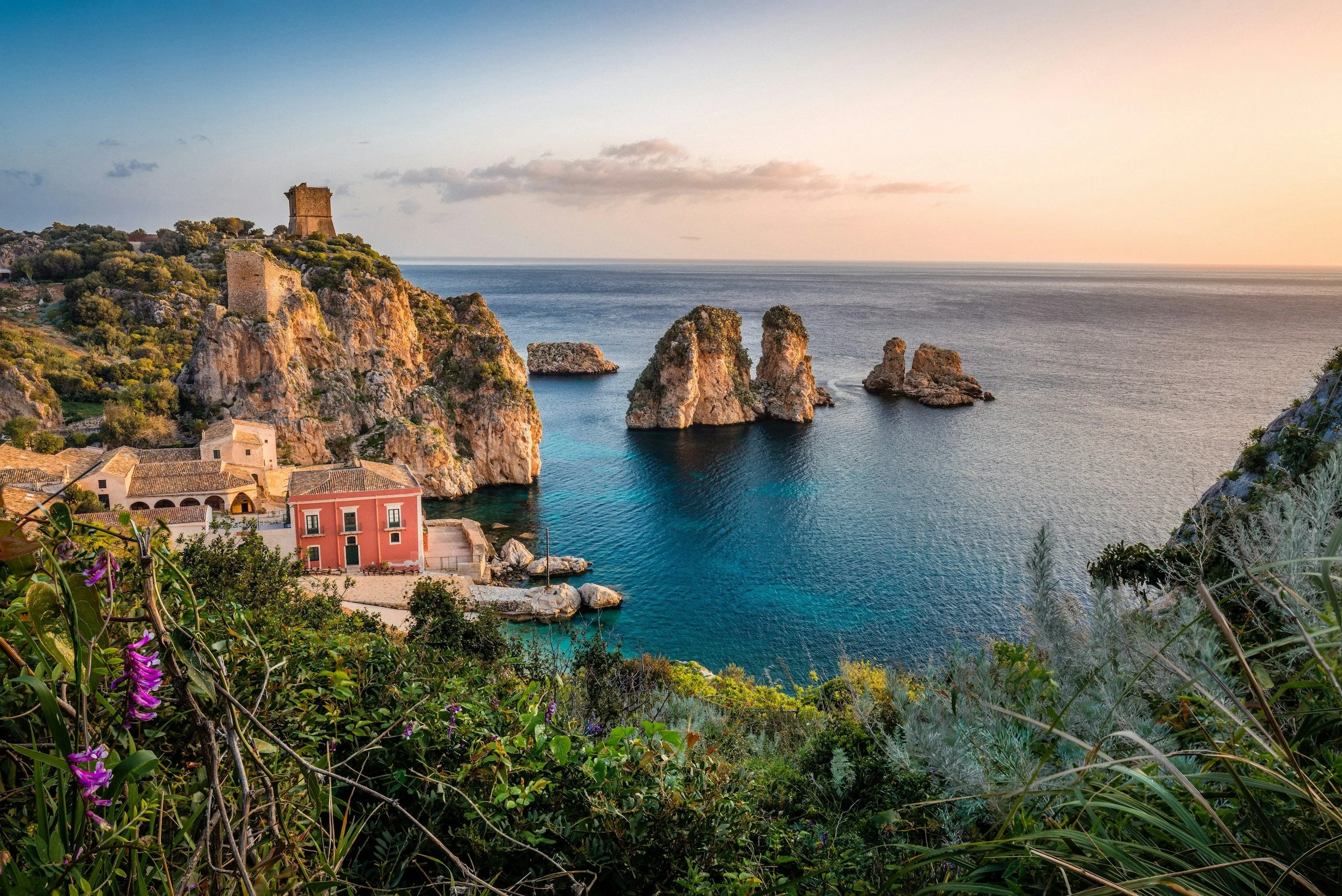 Coastal landscape with rocky cliffs, a small village with pastel-colored buildings, and historic towers overlooking the sea at sunset.